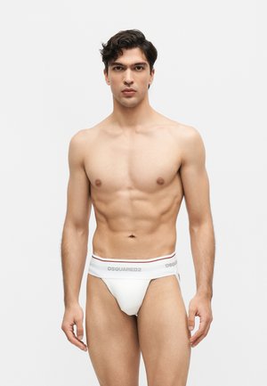 Jeune homme mince aux cheveux foncés portant un slip blanc de la marque DSQUARED2, debout devant un fond clair uni, faisant face à l'avant.