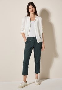 Witte button-up shirt over een witte tanktop, gecombineerd met donkergroene cropped broek en witte sneakers met subtiele groene accenten.
