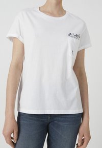 Bílé bavlněné tričko s kulatým výstřihem a ohrnutými rukávy. Má malou kapsu s květinovou výšivkou. Viditelný pár modrých džínů.