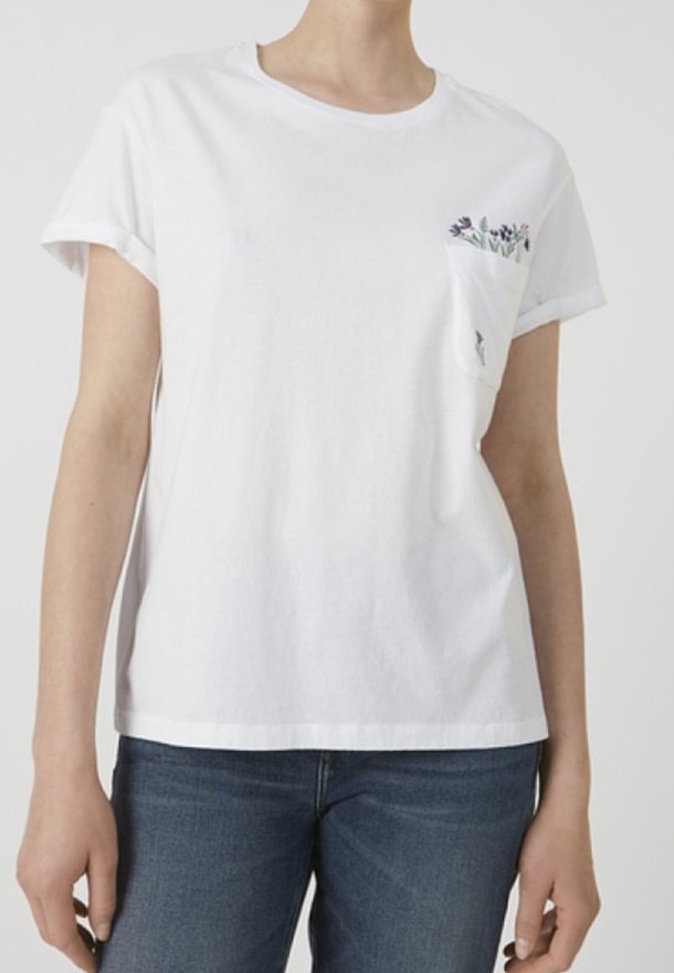 Bílé bavlněné tričko s kulatým výstřihem a ohrnutými rukávy. Má malou kapsu s květinovou výšivkou. Viditelný pár modrých džínů.