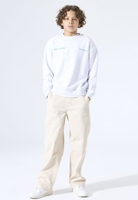 Sudadera gris con texto en verde azulado, corte holgado, escote redondo y puños acanalados. Combinada con pantalones beige y zapatillas blancas.