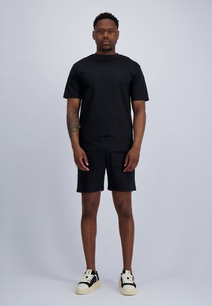 Mann steht frontal, trägt ein schlichtes schwarzes T-Shirt, schwarze Shorts und weiße Sneakers mit schwarzen Akzenten vor einem einfarbigen Hintergrund.