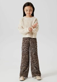 Pull crème à côtes, coupe ample. Pantalons larges à imprimé léopard marron avec une texture lisse. Chaussures beiges à enfiler. Fond neutre.