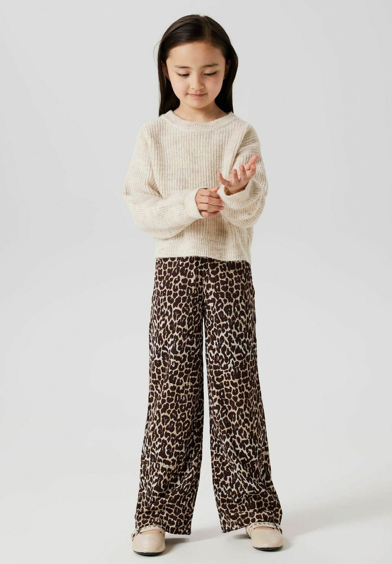 Pull crème à côtes, coupe ample. Pantalons larges à imprimé léopard marron avec une texture lisse. Chaussures beiges à enfiler. Fond neutre.