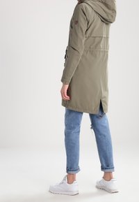 Olivově zelená parka s kapucí, přitažena v pase, se zadními větracími otvory a nastavitelnými šňůrkami, doplněná světle modrými džíny s ohrnutými nohavicemi a bílými teniskami.