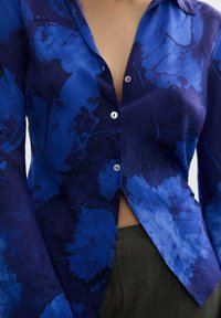 Blaue Bluse mit floralem Muster, die einen tiefen V-Ausschnitt, Knopfverschlüsse und lange Ärmel aufweist. Der Stoff ist glatt und hat ein auffälliges, abstraktes Blumenmuster.