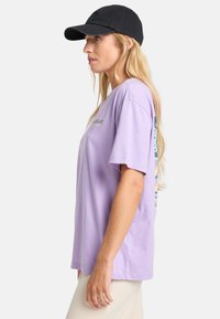 Lilla kortærmet t-shirt med afslappet pasform, der har et logo på forsiden og farverige designs langs siden. Lavet af bomuldsmateriale.