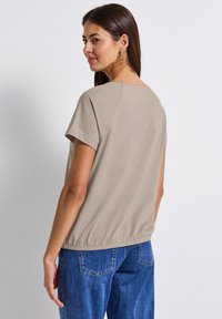 Top beige à manches courtes avec une coupe décontractée et un ourlet élastique. Texture lisse avec des coutures décoratives aux épaules. Assortie à un jean en denim bleu.