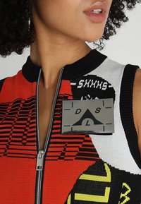 Haut sans manches en tricot avec des motifs rouges, noirs et blancs, un devant zippé et un badge métallique proéminent avec un design géométrique.