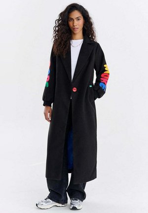 Kobieta o długich kręconych włosach, ubrana w długi czarny płaszcz z kolorowymi literami na rękawach, białą koszulę, szerokie czarne spodnie i srebrne trampki.