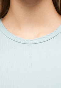 Εγγύτατη λήψη λαιμού και άνω μέρους στήθους που φοράει ένα ανοιχτό μπλε ριμπ πλεκτό πουλόβερ με στρογγυλή λαιμόκοψη.