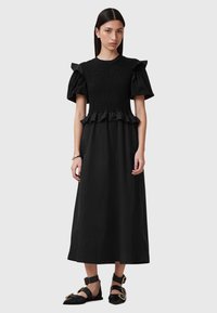 Zwarte jurk met korte pofmouwen, smocktop, gerusht tailleband en een lange rok tot op de vloer. Draagt met platte zwarte schoenen met gouden accenten.