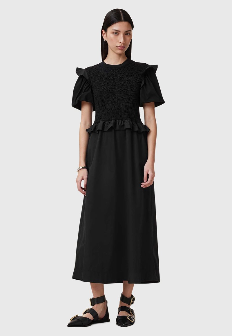 Zwarte jurk met korte pofmouwen, smocktop, gerusht tailleband en een lange rok tot op de vloer. Draagt met platte zwarte schoenen met gouden accenten.