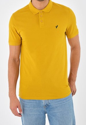 Muž v hořčicově žluté krátkosrsté polokošili se dvěma knoflíky a malým tmavým logem ptáka na hrudi, v kombinaci se světle modrými džínami.