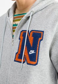 Felpa grigia con zip, dotata di un logo "N" ricamato in texture di colore arancione e blu e un piccolo Swoosh bianco della Nike. Include cordini e tasche laterali.