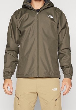 Mężczyzna ubrany w oliwkowo-zieloną, wodoodporną kurtkę na zamek błyskawiczny oraz beżowe spodnie, obie z logo The North Face, stojący na jednolitym tle.