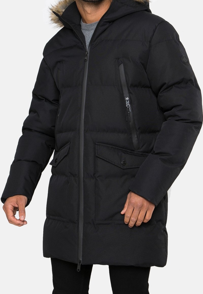 Mann trägt eine schwarze gesteppte Winterjacke mit kapuze mit Fellbesatz, vorderem Reißverschluss, Reißverschlusstaschen auf der Brust und mit Knöpfen versehene Taschenklappen unten.