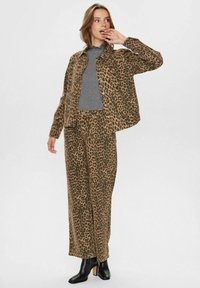 Sako a nohavice so vzorom leopardieho kožucha v hnedej a čiernej farbe, s voľným strihom a hladkým materiálom. Model stojí s rukou pri tvári, obliekaný v čižmách.