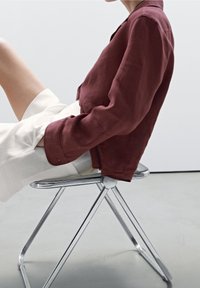 Bordeaux linnen jasje met een kraag en opgerolde mouwen, gecombineerd met crèmekleurige shorts. Het model zit op een zilveren metalen stoel.