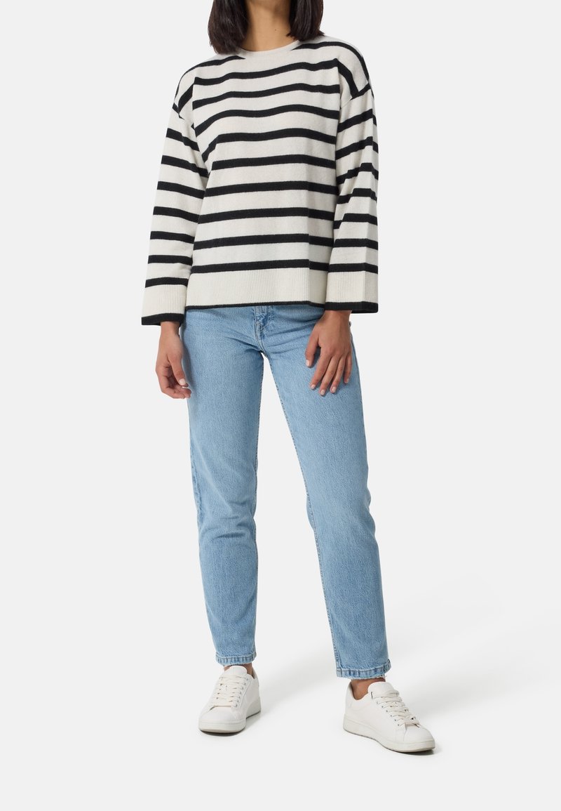 Femme portant un pull à rayures noires et blanches, un jean bleu clair et des baskets blanches, debout devant un fond uni.