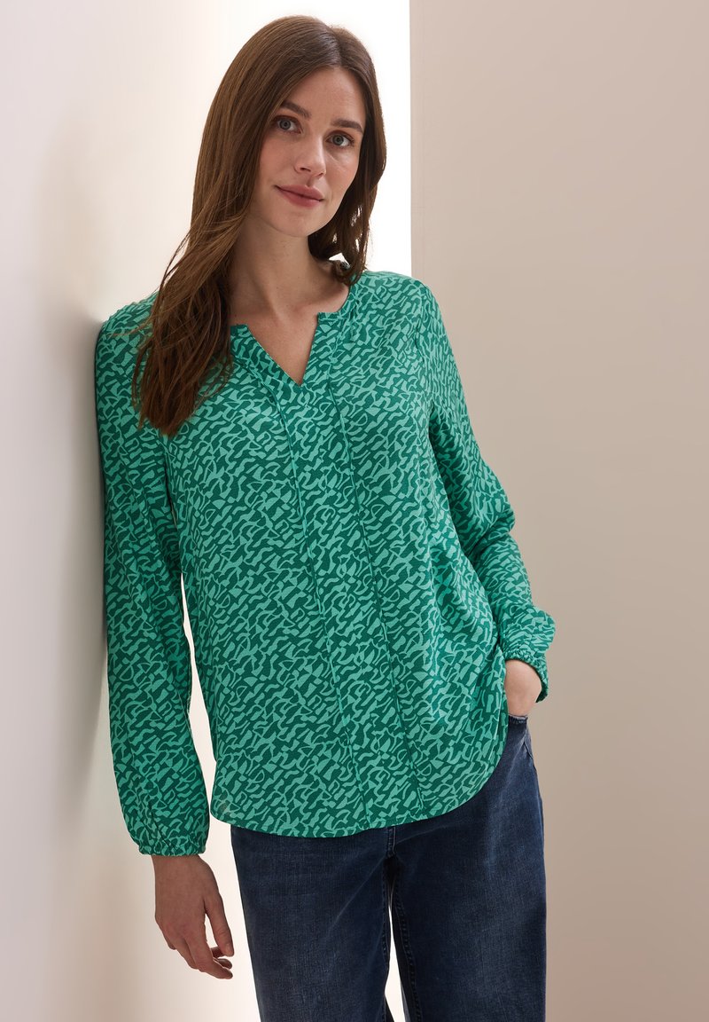 Blouse verte à motif avec un col en V, manches longues avec poignets élastiques, et une coupe décontractée, associée à un jean en denim foncé.