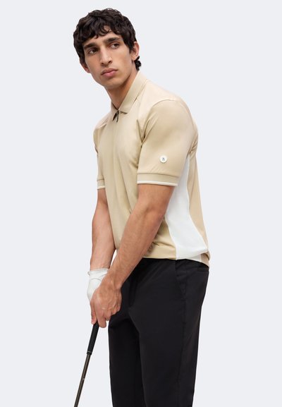 Jeune homme en polo de golf beige et pantalon noir tient un club de golf, se préparant à un swing, regardant vers sa gauche.