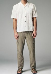 Hvid kortærmet skjorte med lyse striber og brune knapper, kombineret med olivengrønne cargobukser med sidelommer og mørke sandaler.