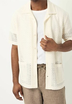 Mężczyzna w kremowej, krótkorękawowej, ażurowej koszuli na guziki noszonej na białym t-shircie oraz beżowych spodniach z wiązaniem w pasie, z ręką przy klatce piersiowej.