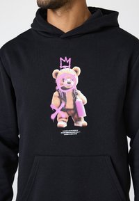 Schwarze Kapuzenjacke mit einem Grafiken von einem Teddybären mit einer lila Krone und einer Sprühdose. Mit pinken Akzenten und einer Kängurutasche.