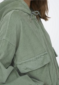 Veste en velours côtelé vert clair avec capuche, dotée d'une fermeture éclair, de deux poches avant et d'un tissu texturé avec des côtes verticales.