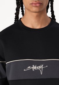 Sudadera negra con una franja horizontal gris, con ribetes blancos y un bordado estilizado del nombre de la marca en el pecho.