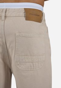 Beige cotton pants featuring a back pocket with embroidered "PEGADOR" logo and a brown leather patch at the waistband. White elastic band visible.