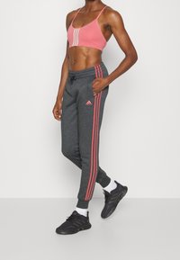 Persona atlética con un sujetador deportivo rosa y pantalones deportivos gris oscuro con rayas rosas, de pie con una mano en el bolsillo contra una pared blanca.