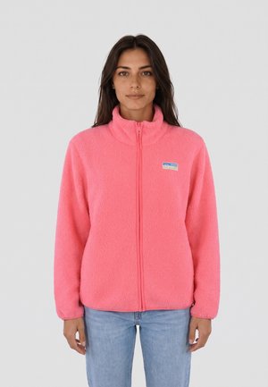 Donna che indossa una giacca in pile rosa acceso con zip frontale e jeans azzurri chiari, in piedi davanti a uno sfondo grigio uniforme.