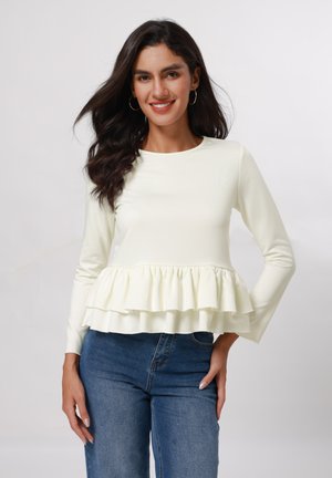 Long-sleeve, light yellow top with a ruffled hem. Smooth texture, fitted design, paired with blue jeans. Simple round neckline.