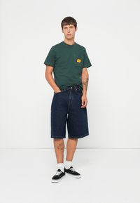 Camiseta de algodón verde con un bolsillo en el pecho que presenta una etiqueta amarilla, combinada con pantalones cortos de mezclilla azul oscuro. El modelo lleva zapatillas negras y calcetines blancos.