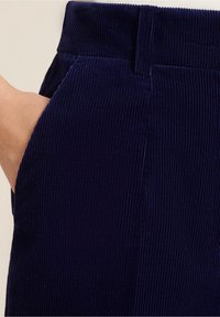 Donkereblauwe corduroy broek met verticale ribbels, voorzien van een zijzak en een tailleband. De textuur lijkt zacht en licht fluweelachtig.