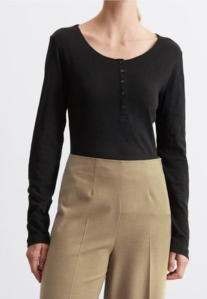 Kobieta ubrana w czarną koszulę henley z długim rękawem, z pięcioma guzikami oraz beżowe spodnie z wysokim stanem na jednolitym tle.