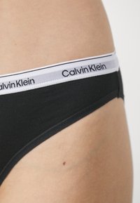 Tanga in cotone nero con una cintura a contrasto bianca riportante il logo "Calvin Klein". Texture liscia, copertura minima e nessun motivo aggiuntivo.