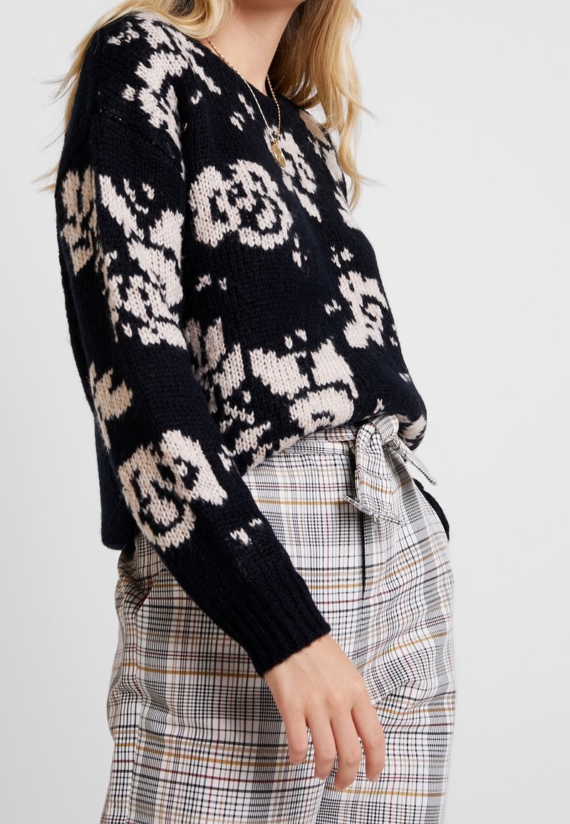 Stickad tröja i marinblått med beiga blommönster, som har en avslappnad passform och ribbade ärmslut. Kombinerad med rutiga hög midja-byxor som är knutna i midjan.