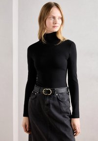 Bluză cu mânecă lungă, neagră, tip guler înalt, combinată cu o fustă din denim negru, cu talie înaltă. Cureaua neagră are o cataramă ovală, aurie, cu un design răsucit.