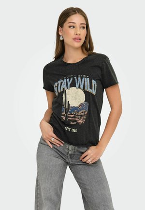 Camiseta gráfica negra con cuello redondo, que presenta una escena del desierto y el texto "MANTENTE SALVAJE". Combinada con unos vaqueros de corte relajado en gris claro.