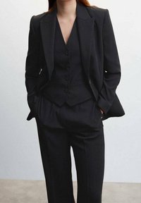 Czarny garnitur szyty na miarę, składający się z dopasowanego blezera z ostrymi klapami, kamizelki na trzy guziki oraz spodni o szerokich nogawkach. Gładka tkanina o wyraźnej, strukturalnej sylwetce.
