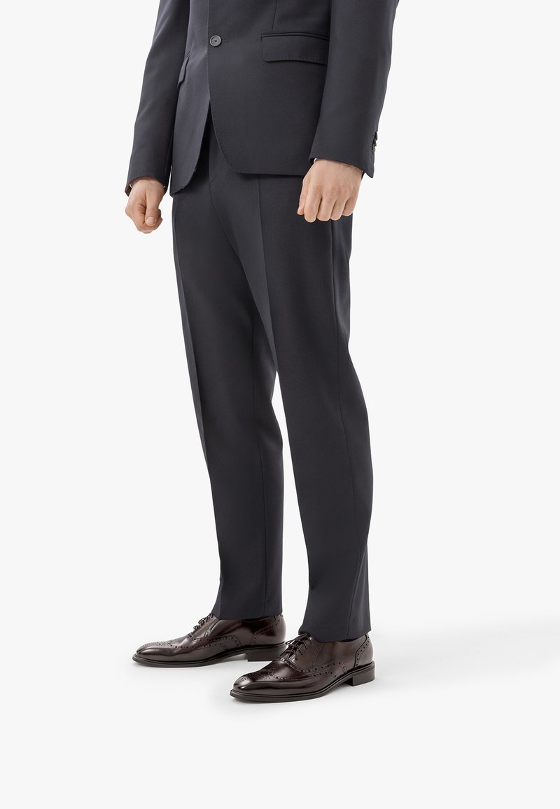 Homme portant un pantalon de costume sombre sur mesure et des chaussures en cuir marron poli, debout sur un fond blanc uni.