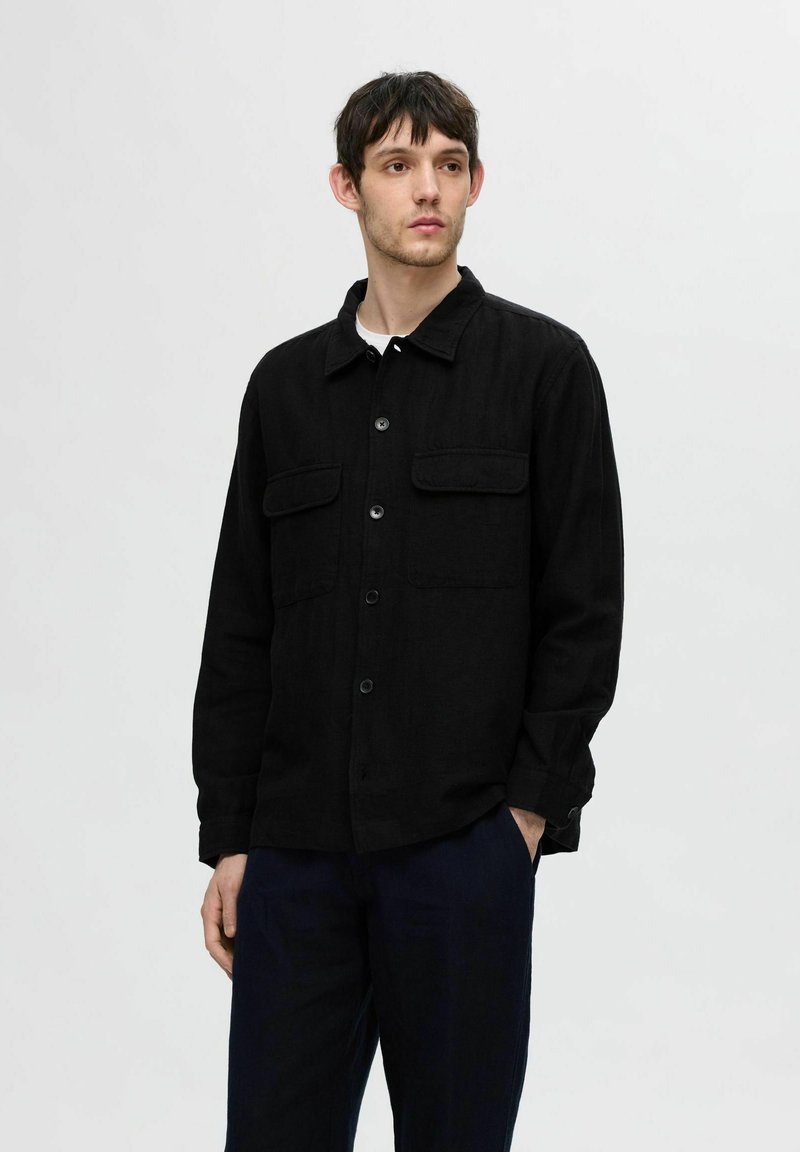 Juoda marškinėliai su apykakle, dviem priekinėmis kišenėmis ir ilgomis rankovėmis. Tekstūruotas audinys, paprastas dizainas, derinami su tamsiomis kelnėmis.