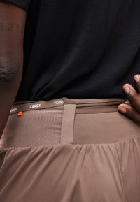 Άτομο που φοράει καφέ παντελόνια Terrex με λεπτομέρεια στη μέση και φερμουάρ στην τσέπη, ντυμένο με μαύρο πουκάμισο, το χέρι ακουμπά κοντά στη μέση του παντελονιού.