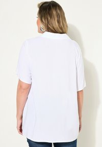 Chemise blanche à manches courtes avec un col, texture lisse et ourlet arrière légèrement plus long, portée avec un jean bleu.