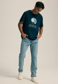 Granatowa bawełniana koszulka z grafiką przedstawiającą ikony tenisa oraz tekst, połączona z jasnoniebieskimi dżinsami i białymi trampkami.