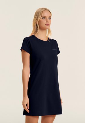 Donna bionda che indossa un vestito nero semplice, a manica corta e lunghezza al ginocchio, con scollo tondo, su uno sfondo chiaro uniforme.