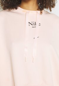Ljusrosa hoodie med avslappnad passform, med en kontrasterande svart Nike-logotyp, dragsko och slät textur.